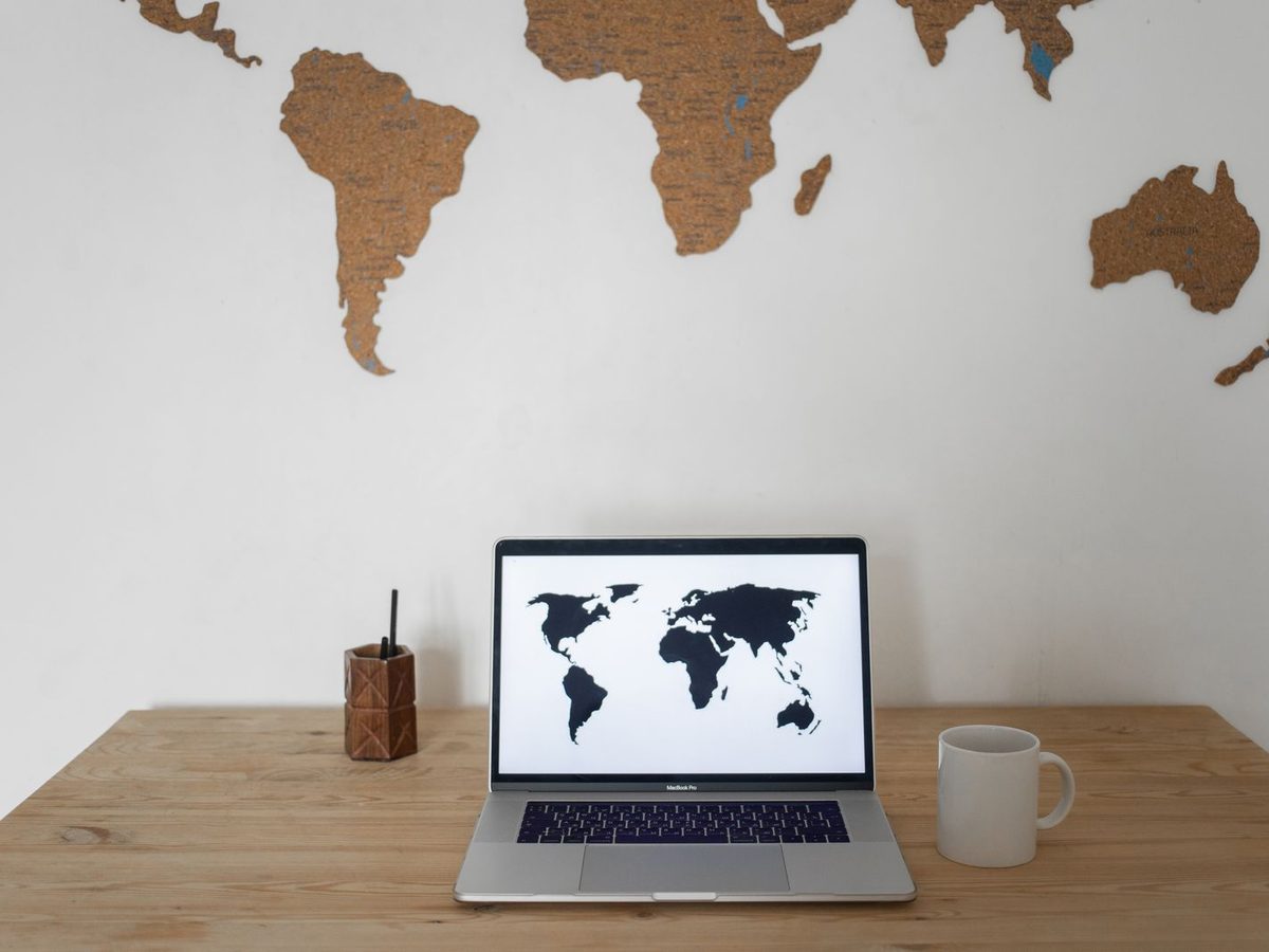A laptop on a wooden desk showing a world map, below a cork world map on the wall, suggesting cross-border selling
